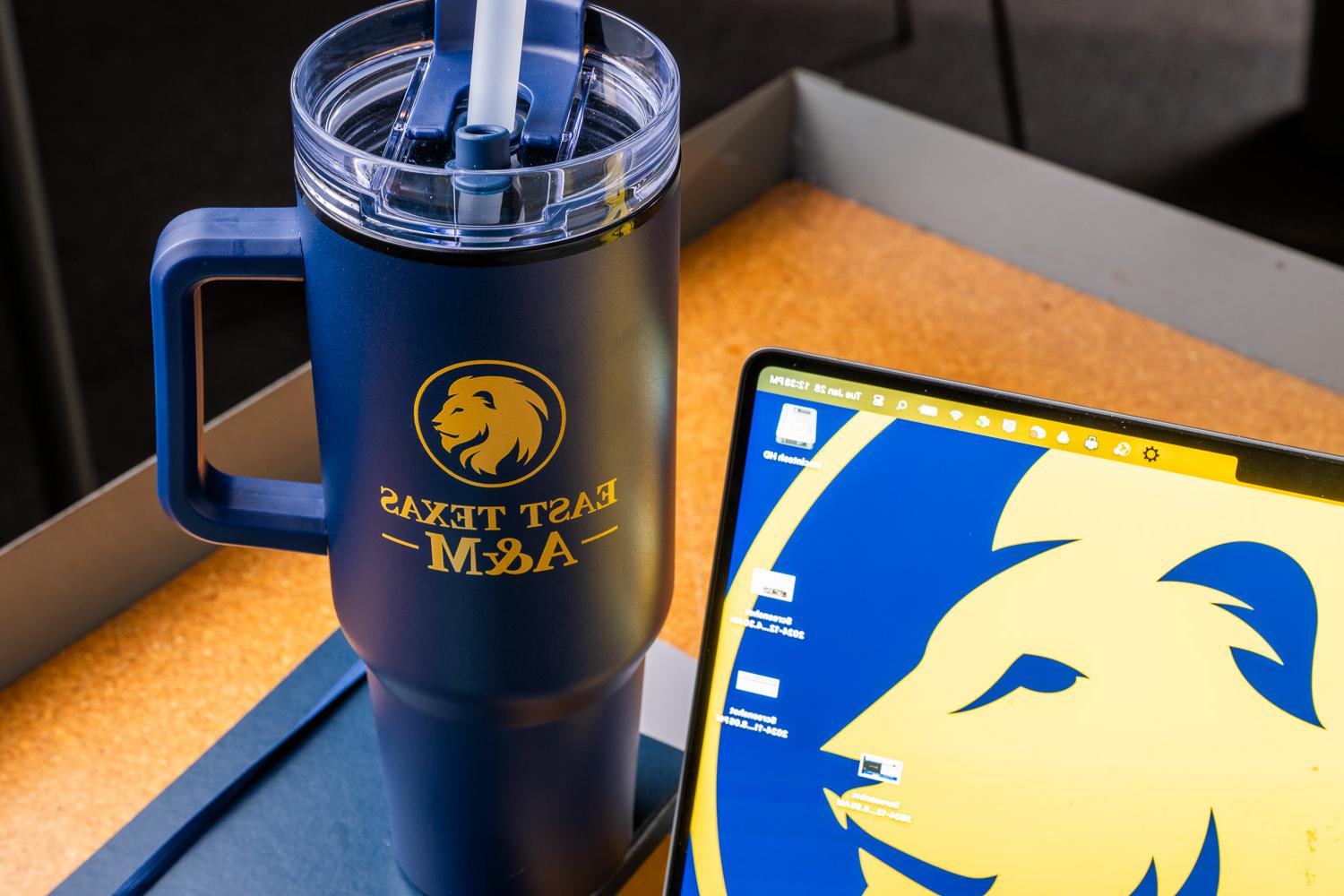 Blue tumbler with East Texas A&M logo and laptop featuring matching lion emblem on screen, photographed on a desk.